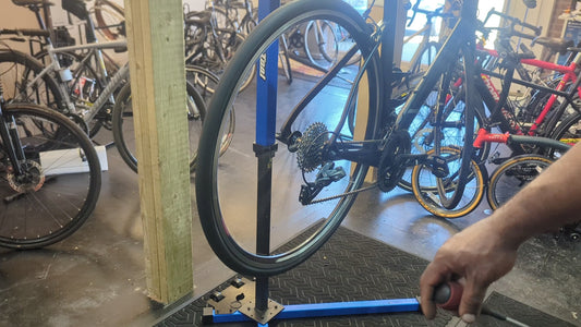 Road bike on a blue workshop stand at Bike Clinique showing the rear wheel, cassette, chain, rear derailleur and crankset, with a hand holding a screwdriver