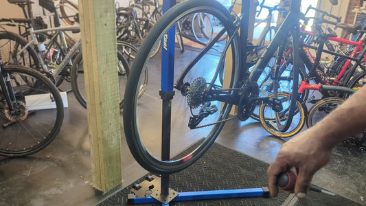 Bicycle rear wheel and drivetrain with cassette, rear derailleur and chain mounted in a blue workshop repair stand, with a mechanic's hand holding a tool