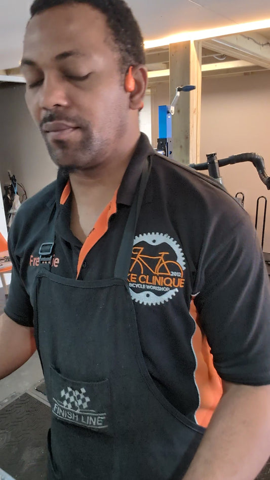 Freddie in a Bike Clinique polo and workshop apron beside the blue repair stand in the Bike Clinique workshop