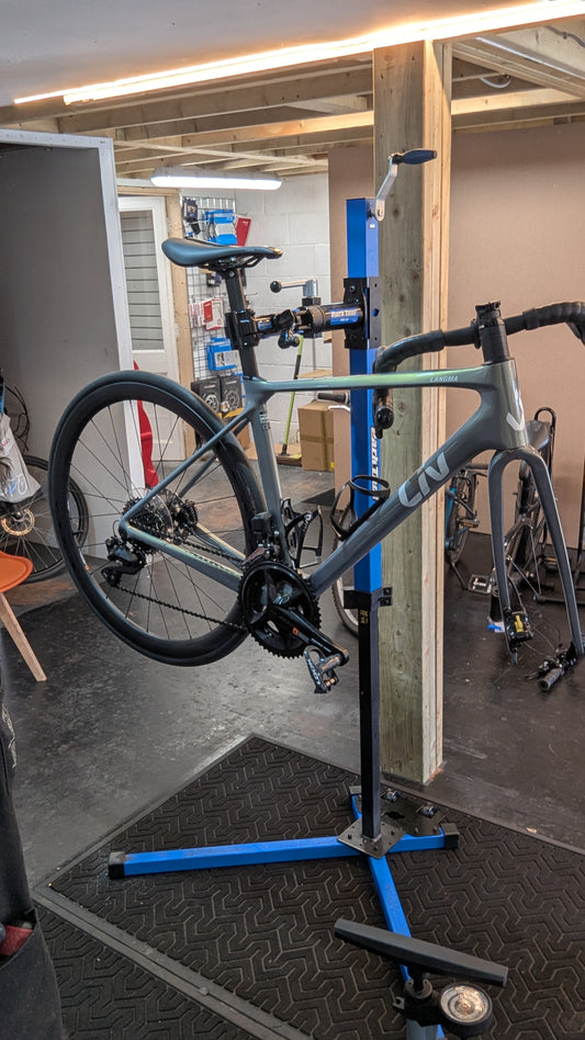 A grey Liv Langma disc brake road bike held in a blue Park Tool workstand at the Bike Clinique workshop with its front wheel removed, showing the bare fork.