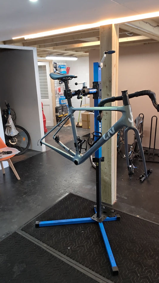 Road bike frame without wheels clamped in a blue repair stand in the Bike Clinique workshop, with handlebar, saddle and fork fitted and loose cables visible