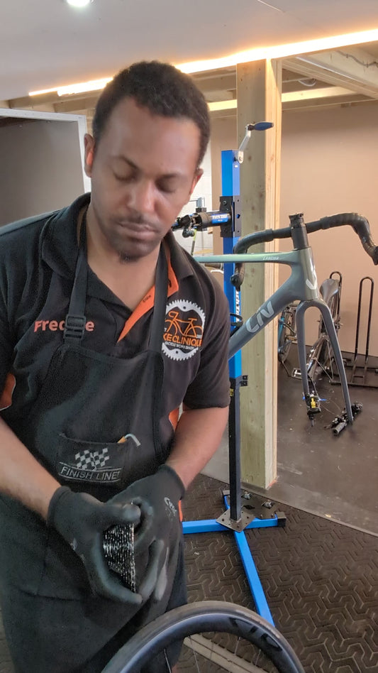 Bike Clinique mechanic in workshop apron holding a rear cassette beside a road bike wheel, with a Liv Langma frame in the repair stand behind