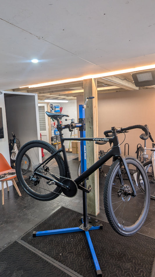 Black drop-bar gravel bike with knobbly tyres and a front disc brake, clamped in a blue workstand in the Bike Clinique workshop