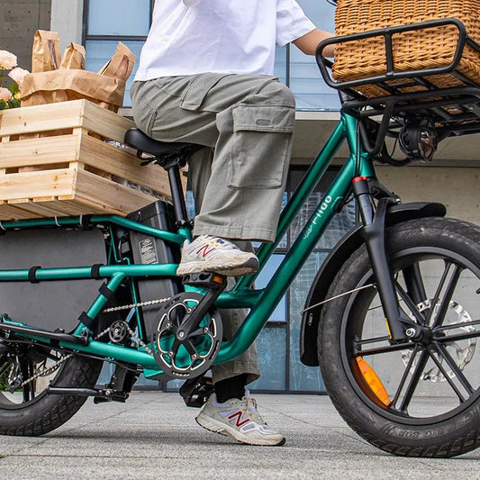 Rider in a white T-shirt and grey cargo trousers seated on a green Fiido T2 step-through longtail cargo e-bike, with a wicker basket on the front rack and a wooden crate of paper bags and flowers on the rear rack