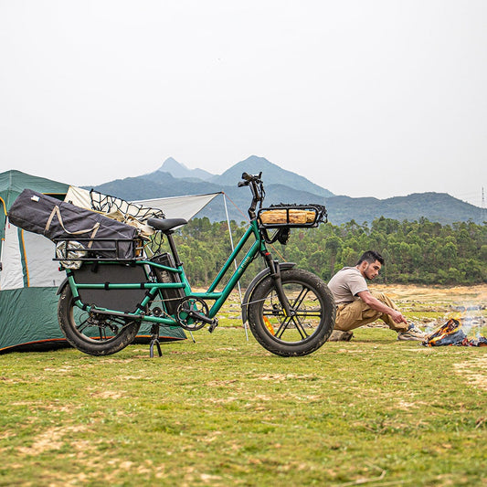 Green Fiido T2 longtail cargo e-bike standing on its kickstand on grass beside a tent, with a bag strapped to the rear rack and a loaded front basket, and a man crouched by a campfire behind