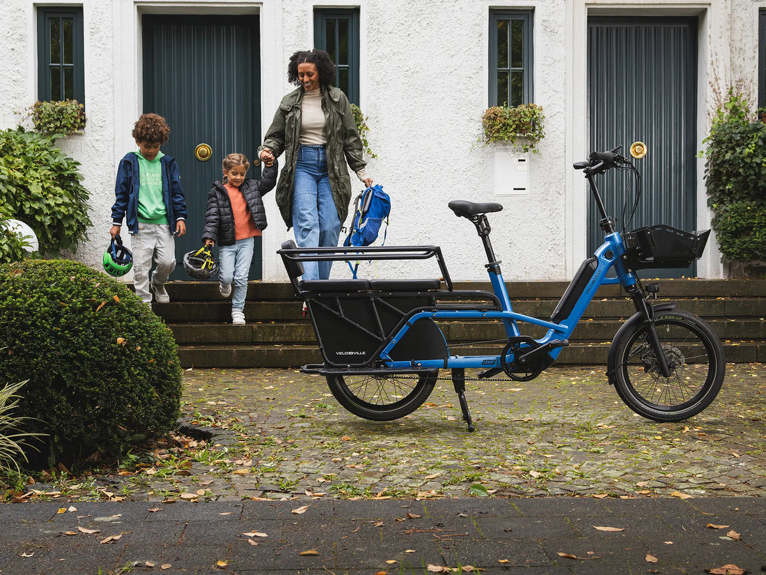 Blue VELO DE VILLE LOADY electric longtail cargo bike with a two-seat rear bench, parked outside a house as an adult and two children carrying cycle helmets and a backpack walk down the front steps