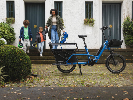 Blue VELO DE VILLE LOADY electric longtail cargo bike with a two-seat rear bench, parked outside a house as an adult and two children carrying cycle helmets and a backpack walk down the front steps